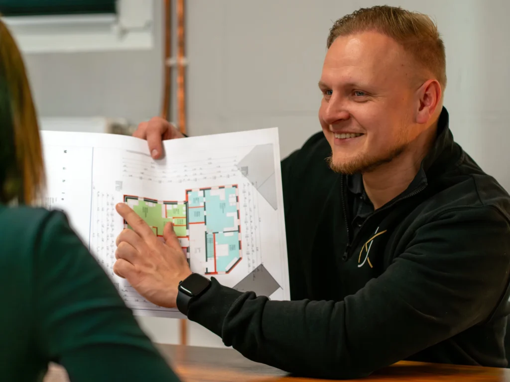 Ein lächelnder Mann zeigt einer Person im Vordergrund einen farbigen Grundriss auf Papier.