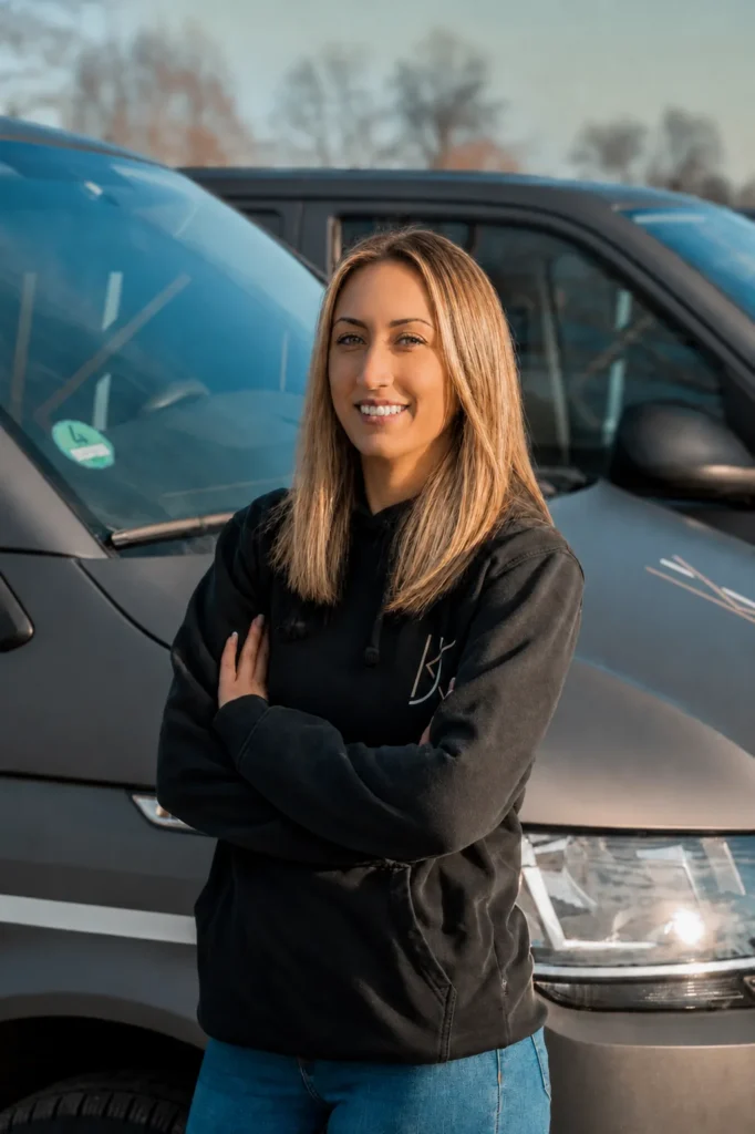 Eine Frau im schwarzen Firmenpullover steht mit verschränkten Armen vor zwei Firmenfahrzeugen der Jung GmbH.