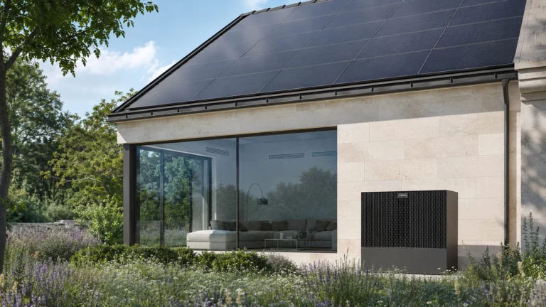 Eine schwarze Wärmepumpe steht vor der hellen Steinwand eines modernen Wohnhauses mit Solardach und bodentiefen Fenstern in einem blühenden Garten.